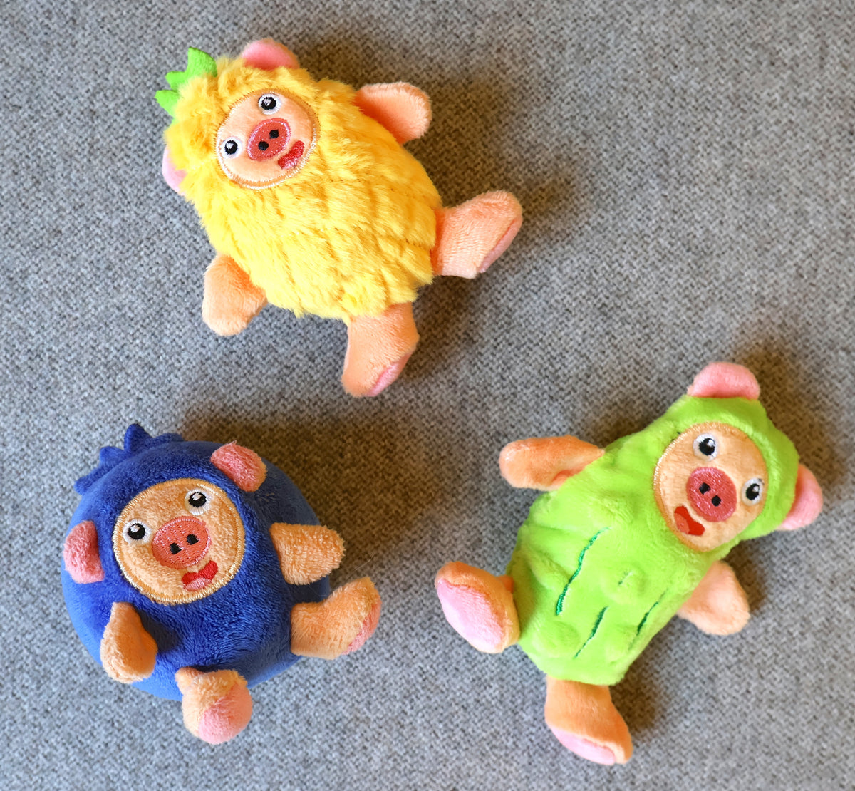 Three small plush toy sheep in different colors (yellow, blue, and green) with pink faces and legs, arranged on a gray textured fabric background.