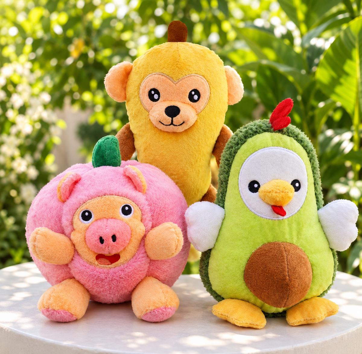 Three plush animal toys arranged on a white table outdoors with a blurred green garden background: a pink pig, a yellow monkey, and a green avocado chick.